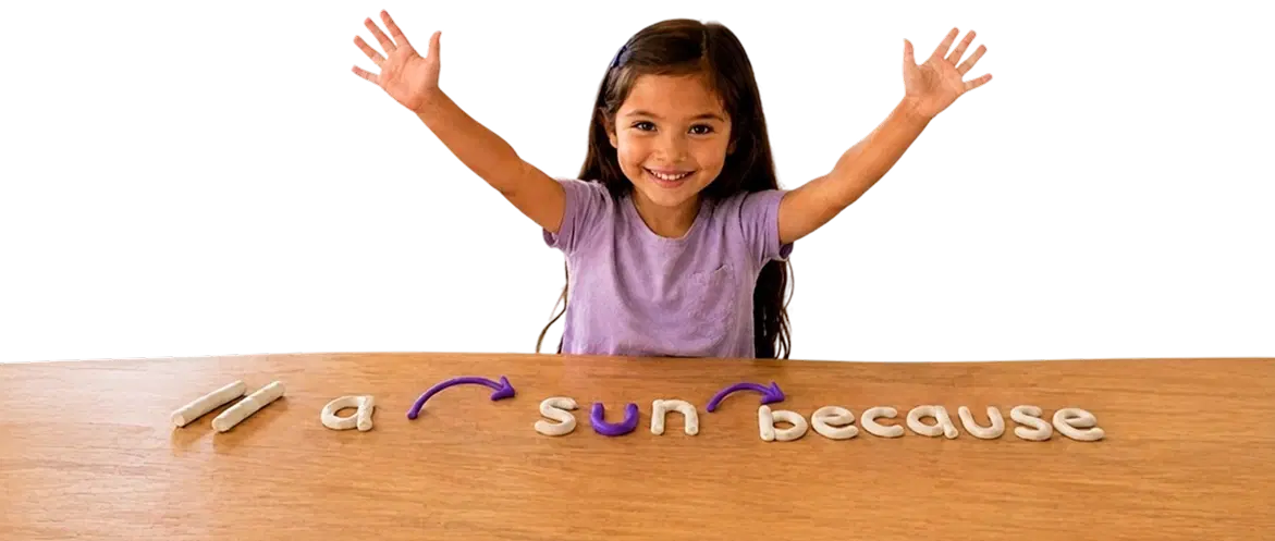 A smiling child raising both hands over a table with clay letters spelling reading practice words