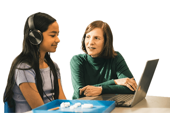 A student wearing headphones smiling at a teacher seated at a laptop, with clay-modeling supplies on the desk between them