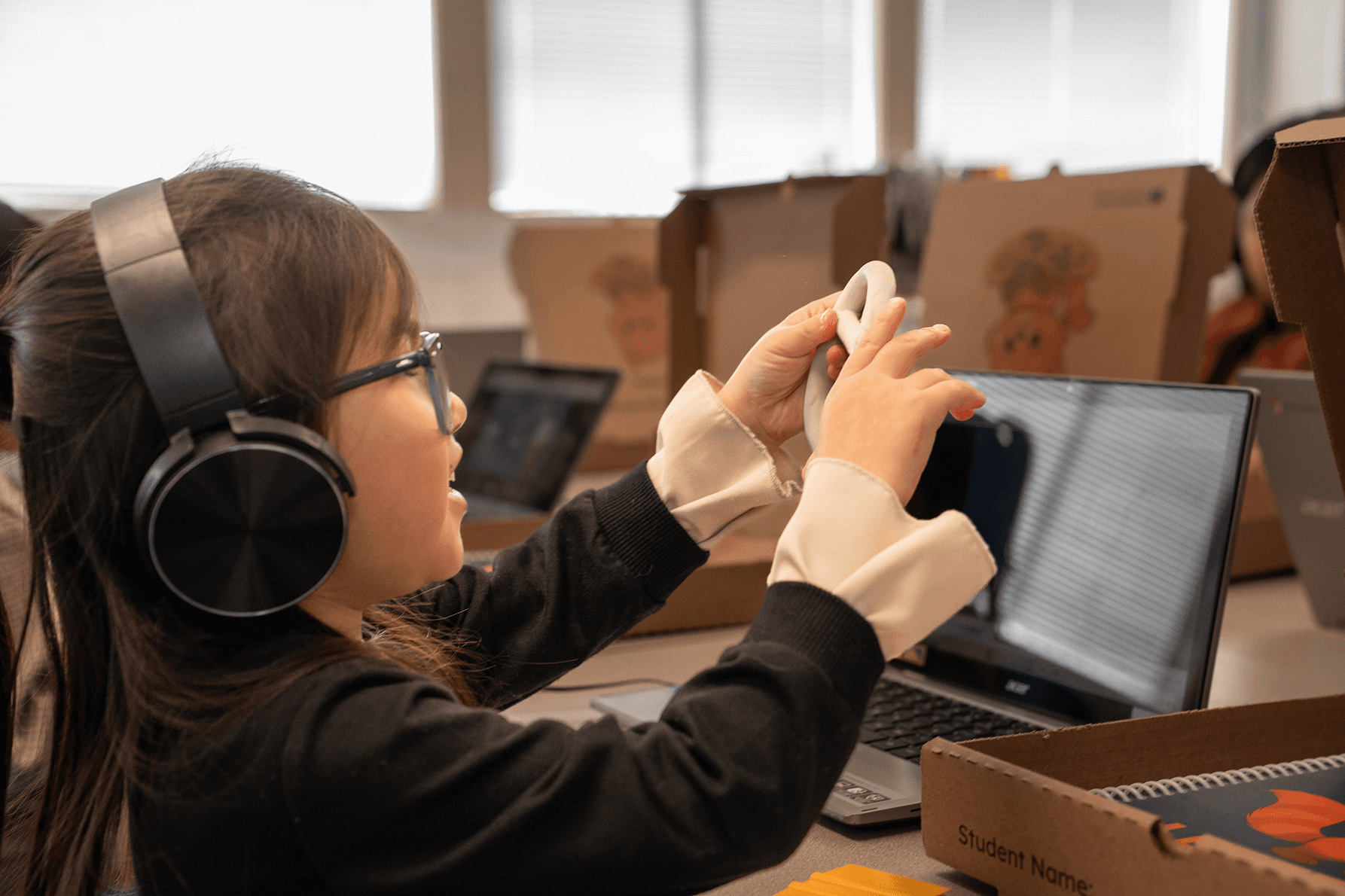 A student wearing headphones shapes a piece of clay while working at a laptop, with a BrainScience Works materials box on the desk